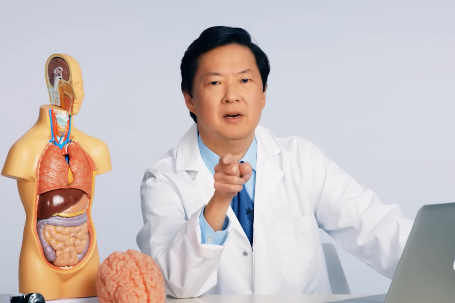 A person in a lab coat points at the camera while sitting next to a human anatomy model and a brain model. The setting is serious and educational.
