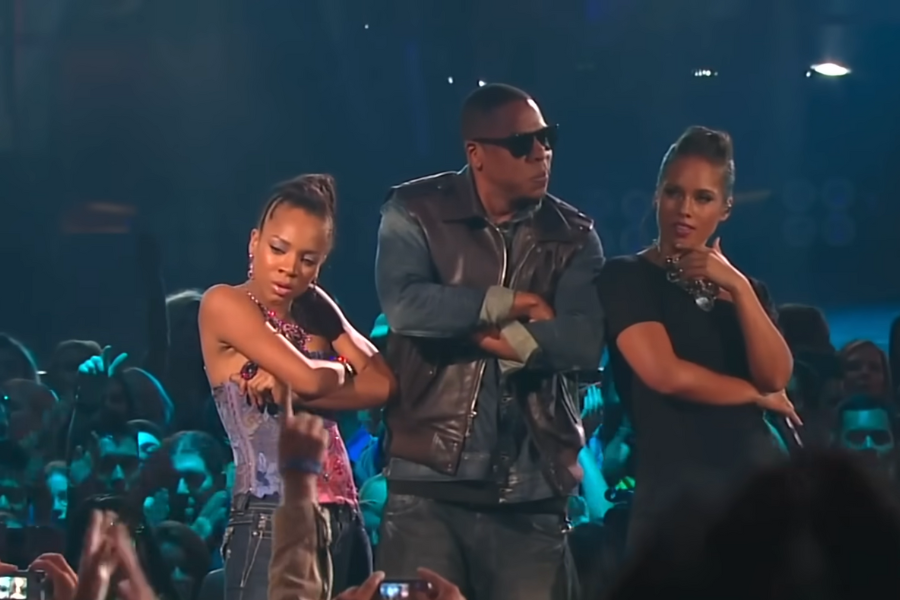 Three people stand on stage with arms crossed, exuding confidence. The atmosphere is lively, with a crowd in the background and blue lighting.