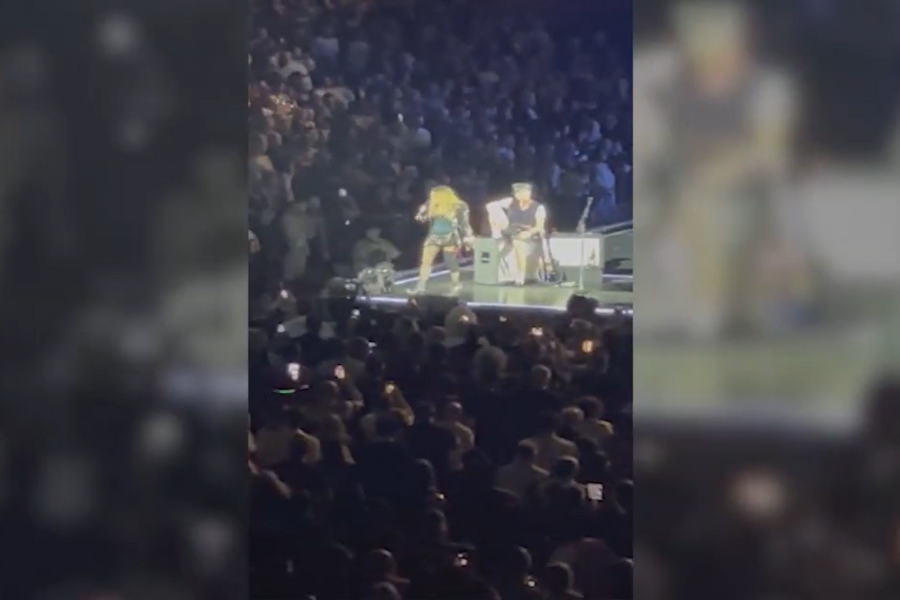 A performer with long hair on stage, possibly singing or dancing, beside a seated individual with a guitar. They are surrounded by a large, attentive crowd.