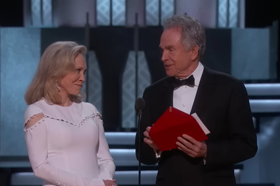 An elegantly dressed woman and man stand on stage, exchanging glances with slight smiles. The man holds a red envelope, suggesting an award event.