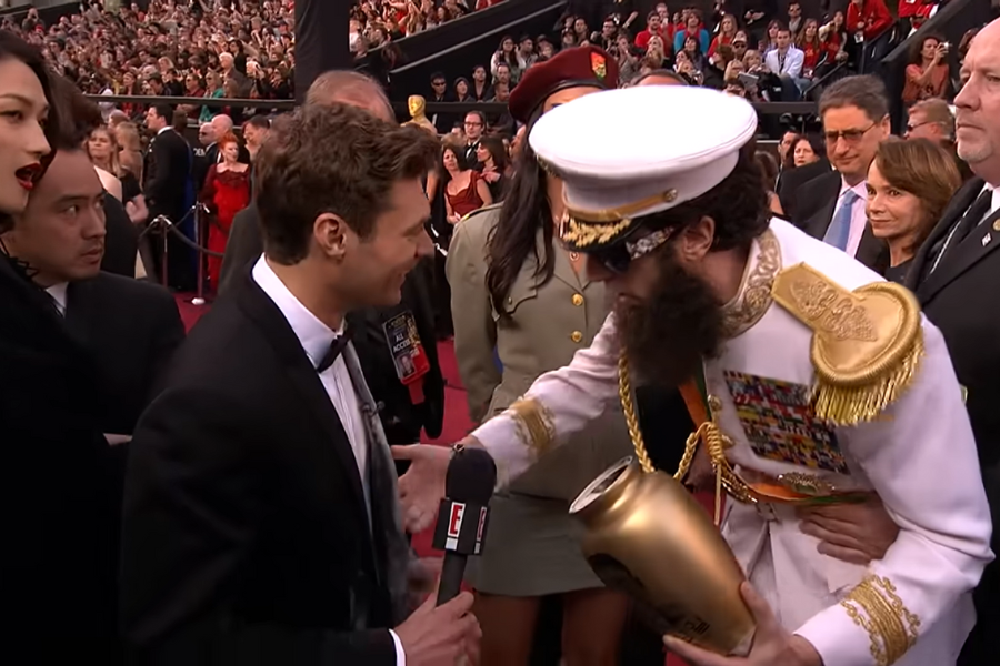 A man in a tuxedo interviews a bearded person in a decorated military outfit holding an urn on a crowded red carpet. The scene is lively and humorous.