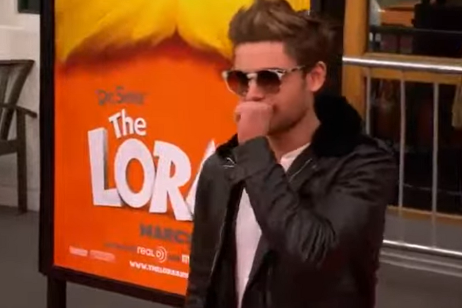 A person in sunglasses and a leather jacket covers their mouth, standing near a poster for "The Lorax." The scene conveys a casual, thoughtful mood.