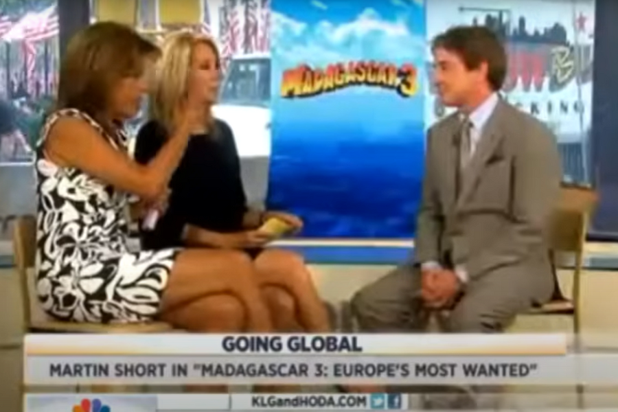 Two women sit on a studio set interviewing a man in a suit. Behind them is a poster for "Madagascar 3: Europe's Most Wanted." The mood is lively.
