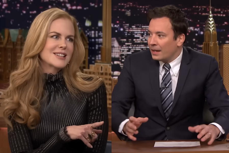 TV talk show scene with a woman in a black dress and a man in a suit. They appear engaged in animated conversation, set against a cityscape backdrop.