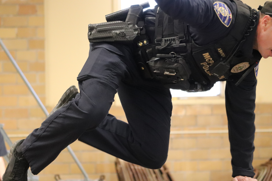 A police officer in tactical gear mid-action while climbing or vaulting over an obstacle indoors during training.