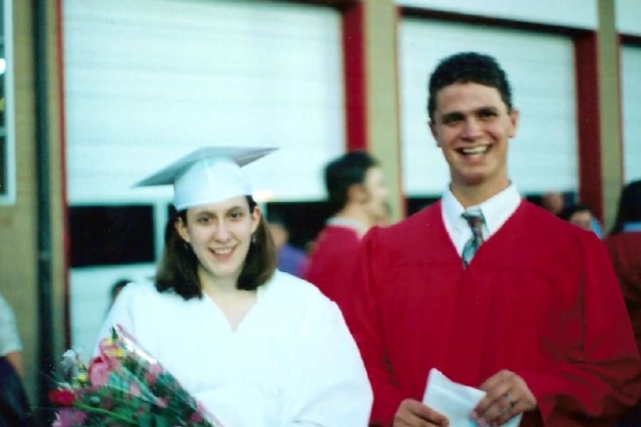 A young woman in a white graduation gown and cap holds a bouquet of flowers while standing next to a smiling young man in a red gown and tie. They appear to be at a high school or college graduation event.