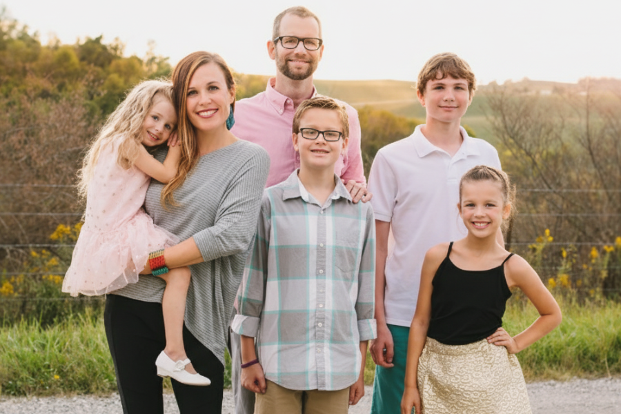 A smiling family of six poses outdoors in front of a wire fence and rolling hills during golden hour.