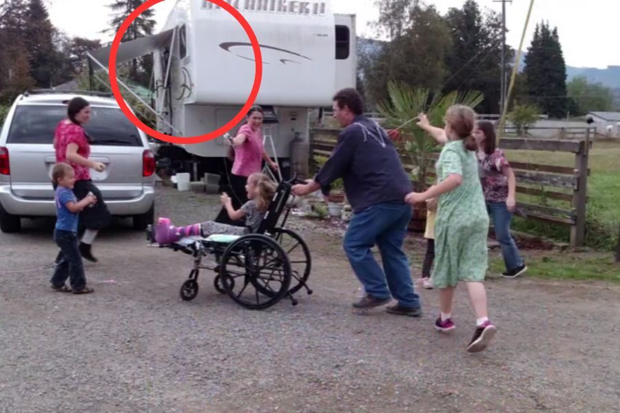 A man playfully pushes a smiling child in a wheelchair while other children and adults engage around them in a gravel driveway with a large white RV in the background.