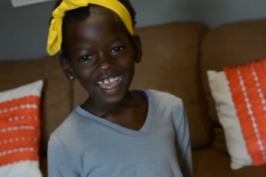 A young girl smiling brightly while wearing a yellow headband and gray shirt in a living room with orange-accented pillows.
