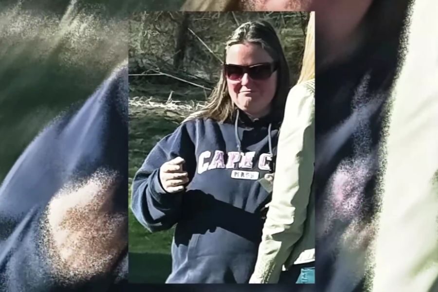 A woman wearing sunglasses and a navy hoodie with “Cape Cod” text stands outdoors, holding a small item in her hand. She is partially turned toward someone beside her.