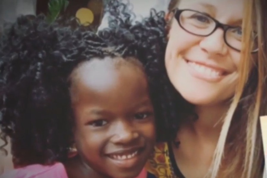 A smiling woman with glasses and a young girl with curly hair pose closely together for a photo.