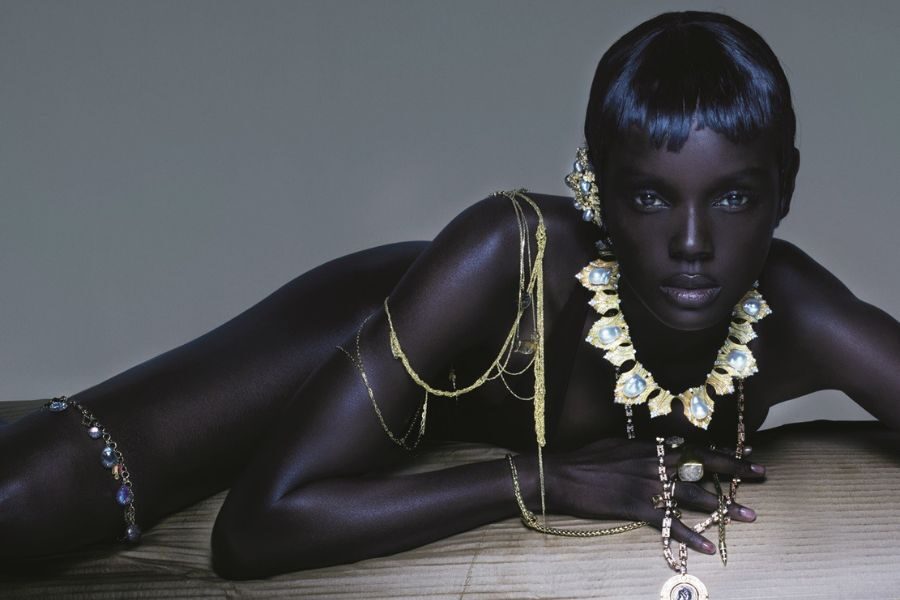 A model with deep skin tone lies adorned in layered gold and gemstone jewelry, styled in a high-fashion, editorial photoshoot.