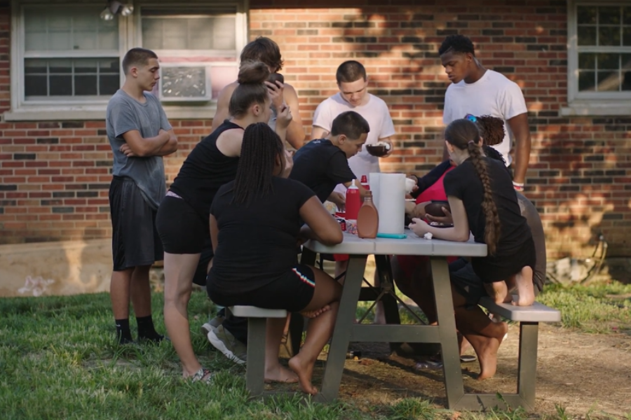 A group of teens and children gathered around an outdoor picnic table in a backyard, enjoying food and conversation.