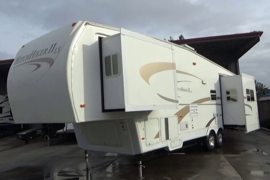 A large white fifth-wheel RV labeled “HitchHiker II LS” is parked on a concrete lot with its slide-outs extended. The sky is cloudy.