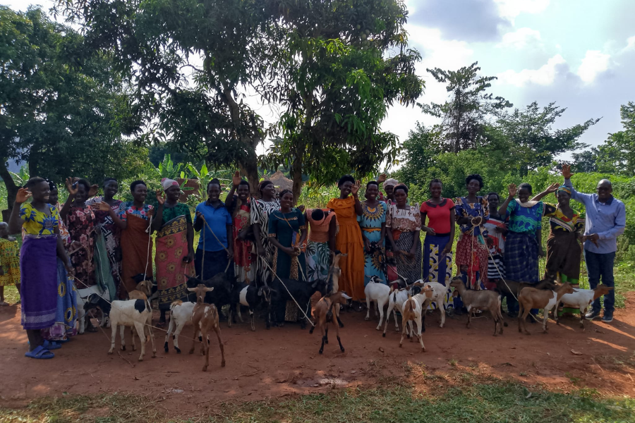 A group of people, mostly women, standing in a row holding goats and waving, with trees and greenery in the background.