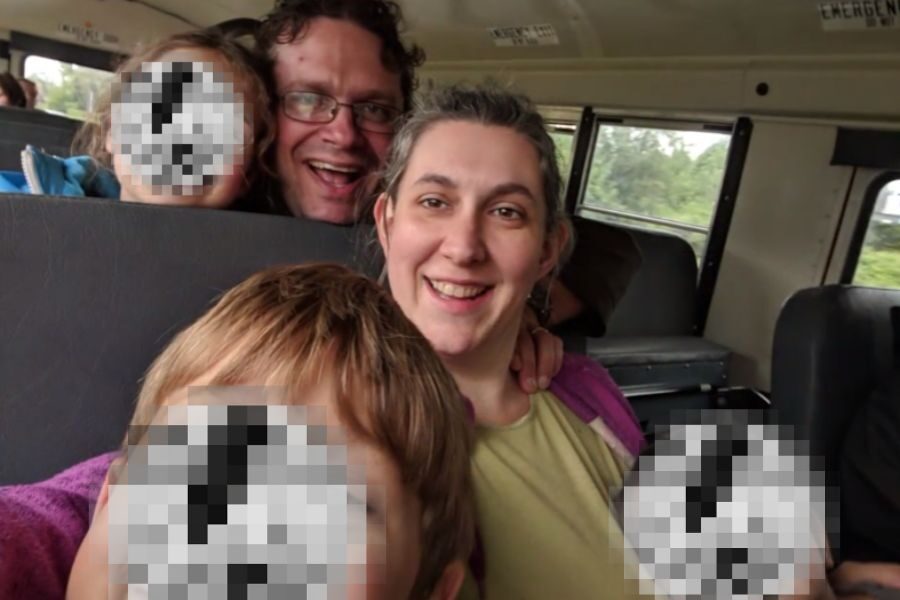 A smiling couple takes a selfie on a school bus surrounded by children with pixelated faces. The woman is seated while the man leans in from behind.
