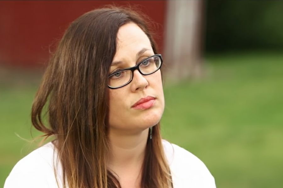 A woman with glasses and long brown hair looks serious while seated outdoors with blurred greenery behind her.