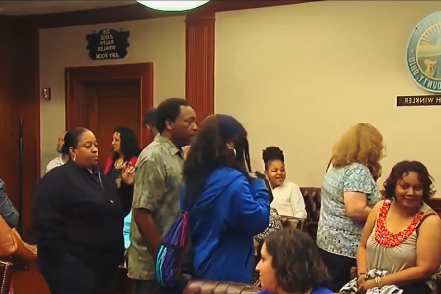 A crowded courtroom scene with adults standing and chatting near the entrance under a judge’s nameplate.