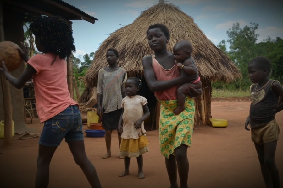A group of children playing near a traditional thatched hut in a rural village, with one girl holding a baby.