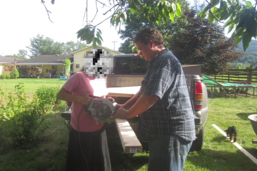 A man and a young person with a pixelated face stand by a truck bed, working together to operate a circular saw on a long piece of wood in a sunny yard.