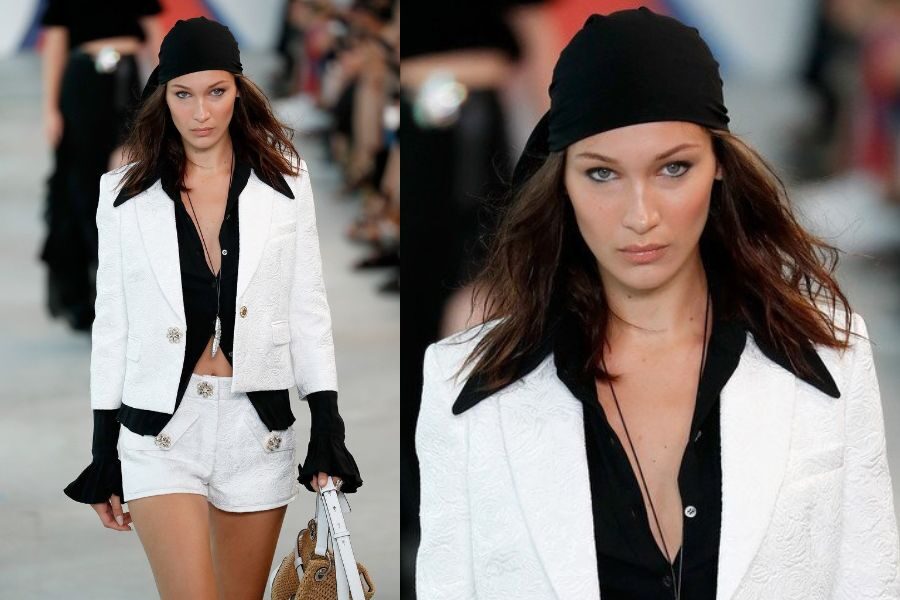 A model on the runway wears a black and white ensemble with gold buttons and a black headscarf, giving fierce eye contact.