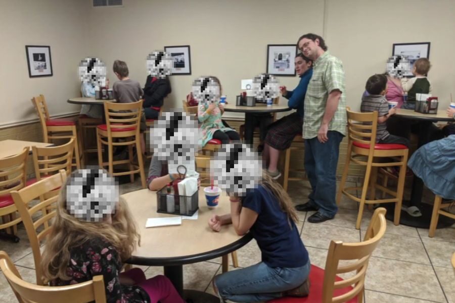 A group of children with pixelated faces sit and stand around tables inside an ice cream shop. Two adults stand near a high-top table smiling at the camera.