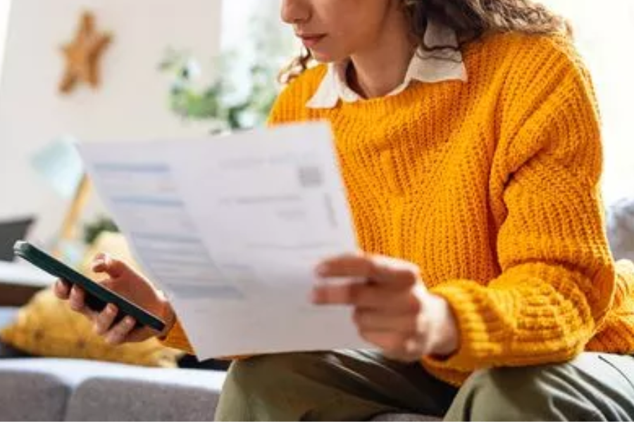A woman in a yellow sweater holds a smartphone and a bill while seated indoors, checking information.