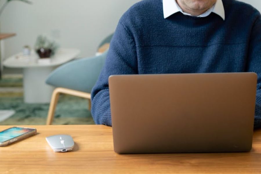 A person in a dark blue sweater works at a wooden desk using a laptop. A smartphone and wireless mouse are placed nearby.