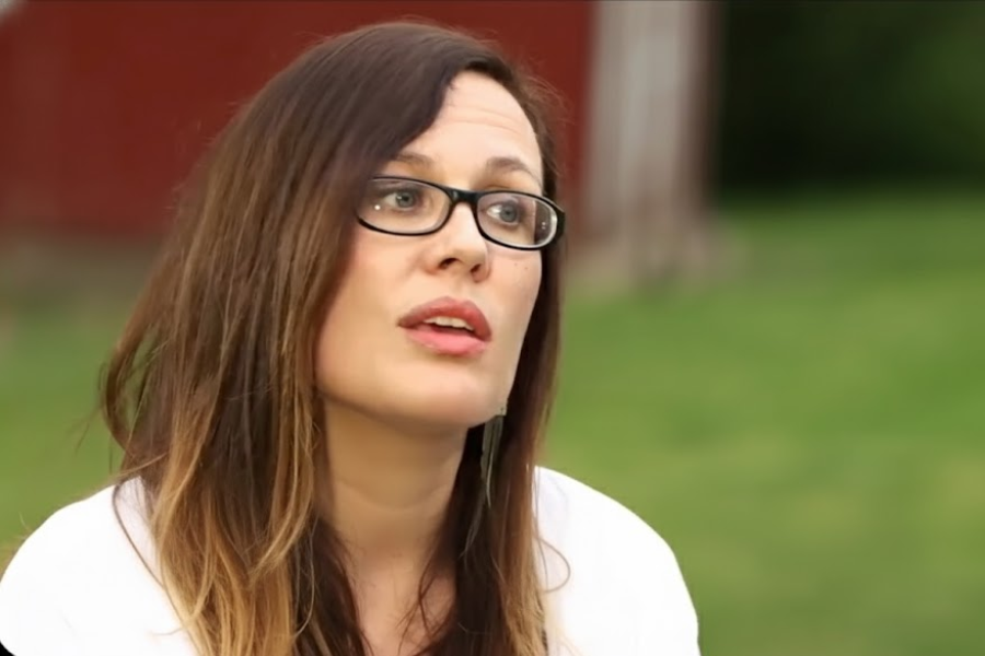 A woman speaking outside with a thoughtful expression, wearing glasses and a white top, with a red building in the background.