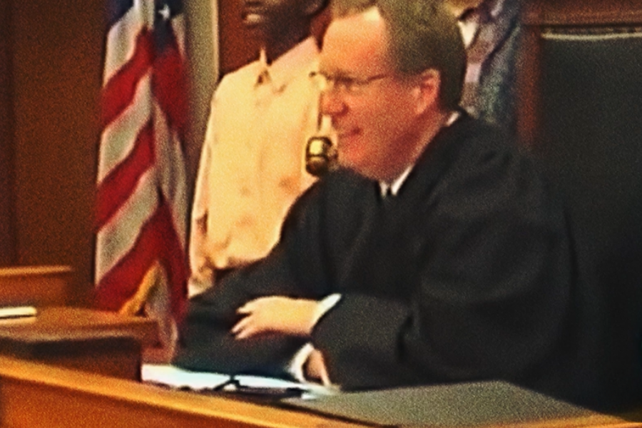A smiling judge sitting at the bench during a court proceeding, with an American flag visible behind him.