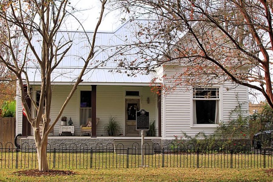 A charming white-painted wooden house with a front porch, gabled roof, and trees with sparse autumn leaves in the foreground.