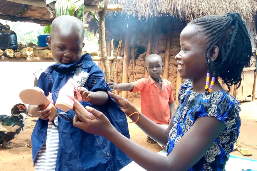A smiling girl gives pink shoes to a baby, with a boy and a chicken visible in a rural setting behind them.