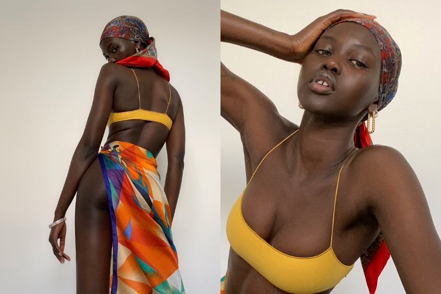 A model in a mustard yellow bikini and colorful headscarf poses with bold energy and a vibrant patterned sarong.