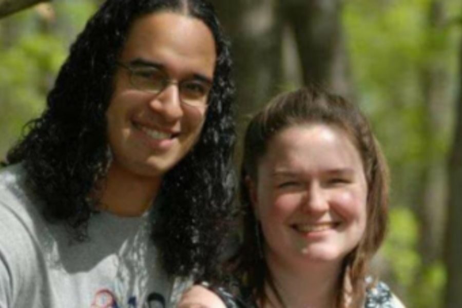 A man and woman smile side by side outdoors with trees in the background. The man wears glasses and has long curly hair, and the woman has her hair pulled back.