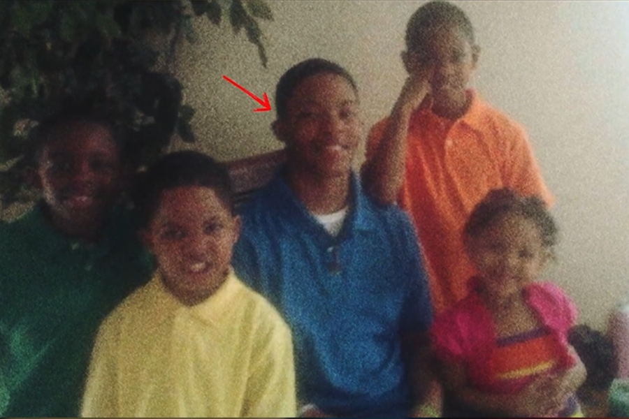 A group of five children smiling for a photo indoors; one boy in a blue shirt is highlighted with a red arrow.