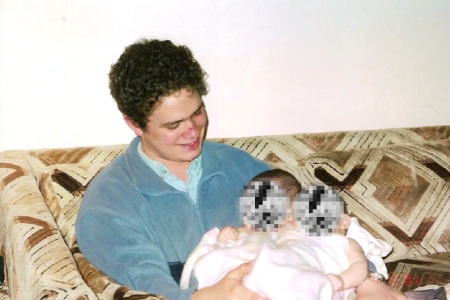 A man in a blue fleece jacket smiles down at two infants wrapped in a white blanket while sitting on a patterned couch. The babies’ faces are pixelated for privacy.