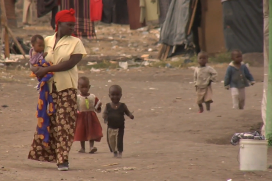 A woman carrying a baby and walking with four young children in a dirt area surrounded by makeshift shelters.