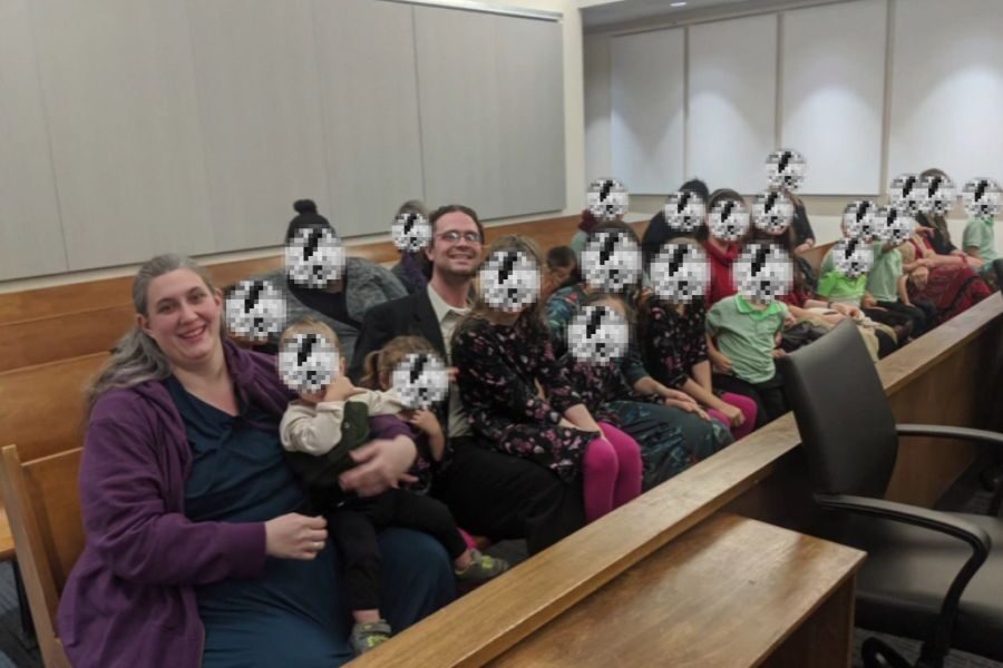 A large family with pixelated children’s faces is seated together in a courtroom pew. The adults smile while surrounded by many children.