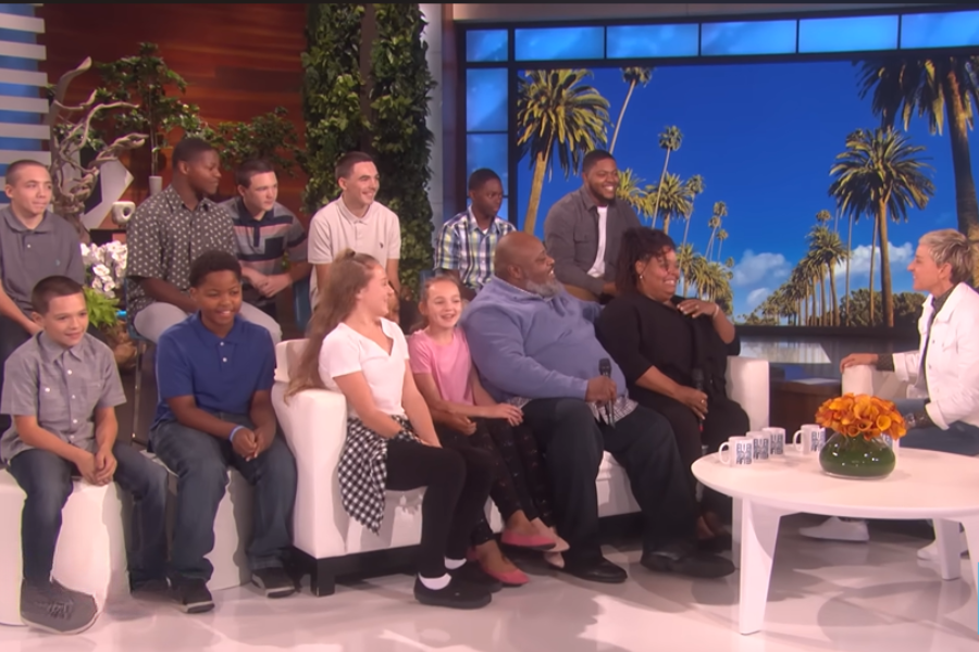 A large family is seated on stage during a talk show, smiling and talking with the host in front of a palm tree backdrop.