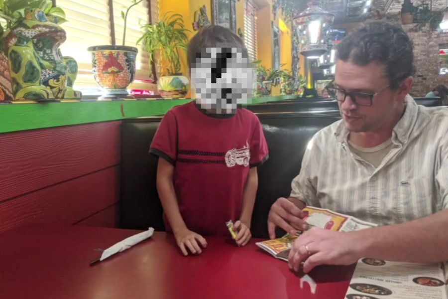 A man and a child sit at a red booth in a colorful restaurant with Mexican-style décor. The man looks down at a kids' menu while the child, whose face is pixelated, stands next to him holding crayons.