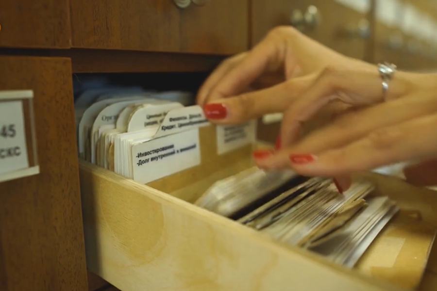 A close-up of a woman’s hands searching through a wooden filing cabinet drawer filled with index cards.