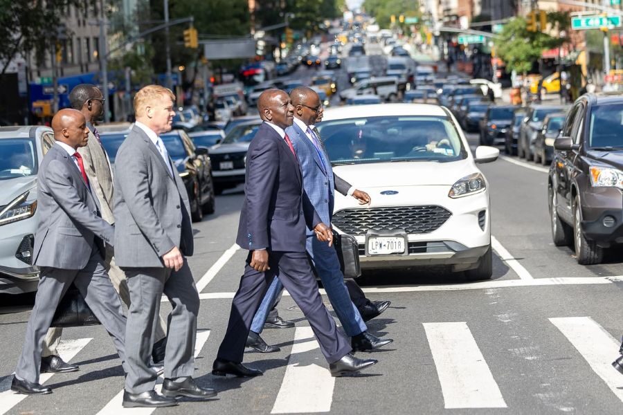 Group of men in suits walking across a busy city crosswalk with traffic stopped behind them.