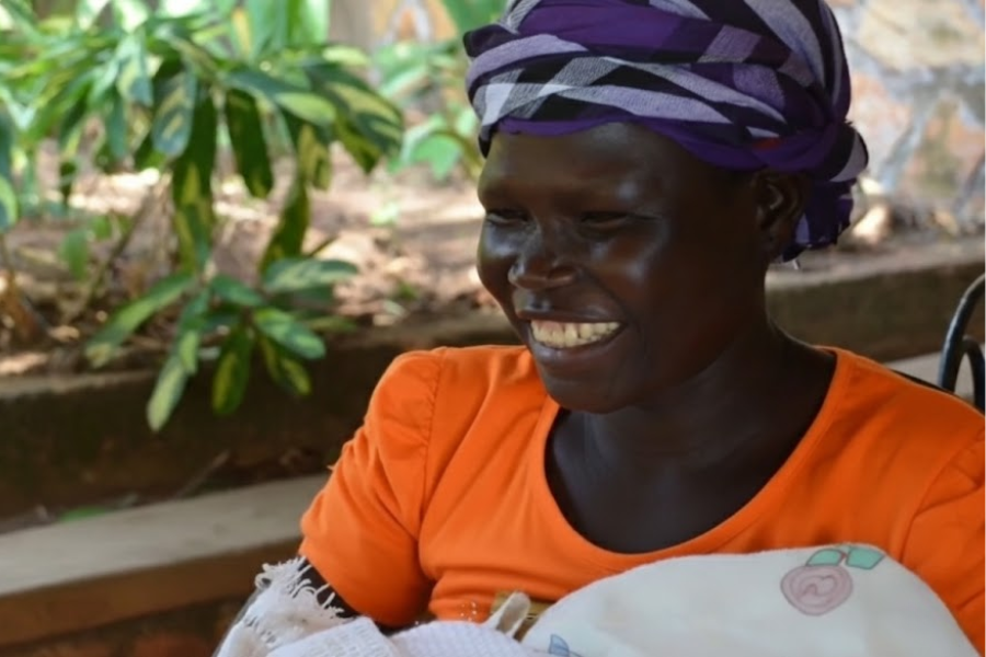 A smiling woman wearing an orange shirt and purple headwrap holds a baby in a blanket outdoors.