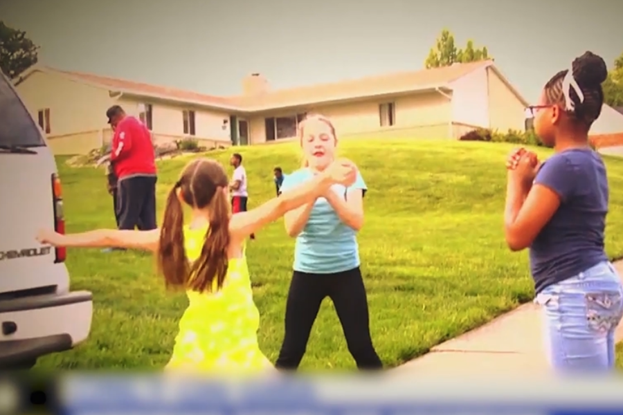 Three young girls play a hand-clapping game on a sidewalk in front of a suburban home, while other children and adults are visible in the background.