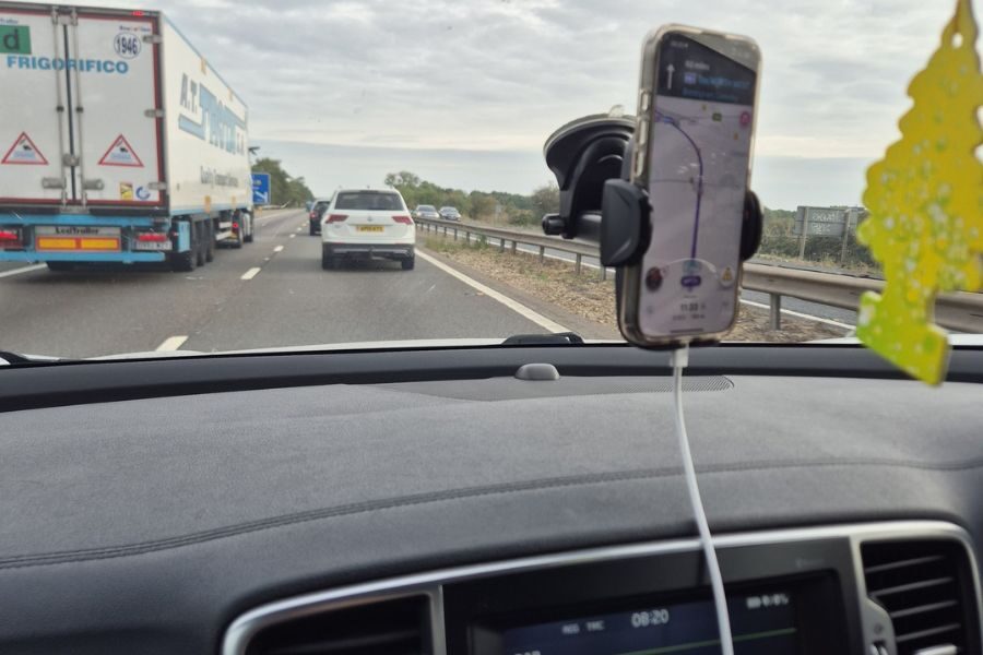 View from inside a moving car showing a smartphone with navigation app mounted on the dashboard while driving on a highway.