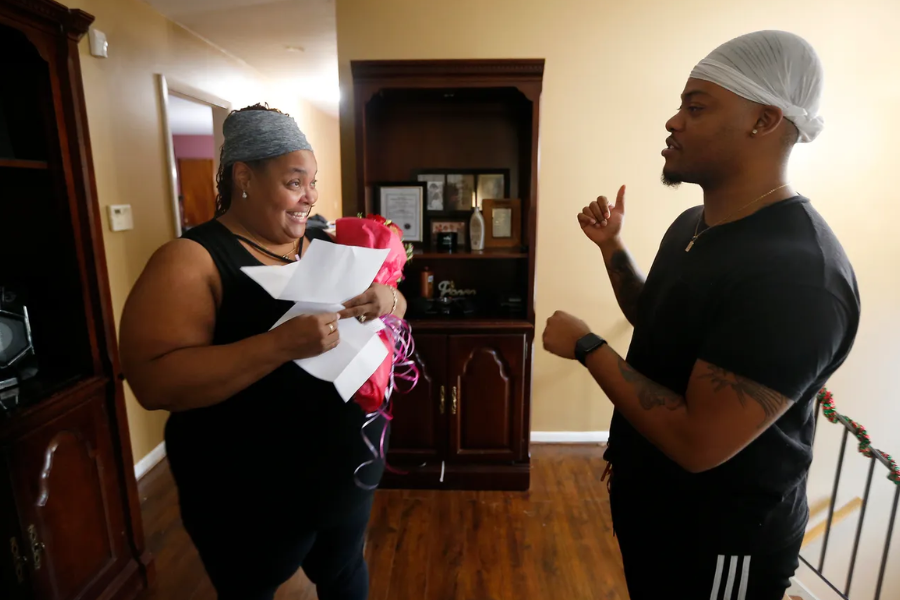 A woman holding papers and flowers smiles at a man speaking to her inside a warmly lit home.