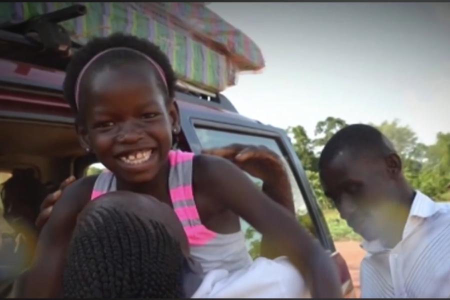 A joyful girl with a pink headband and striped dress smiles while being lifted out of a car by two adults.
