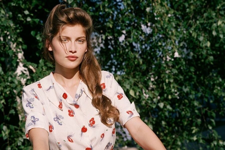 A model in a cherry-print blouse poses outdoors in natural light, her hair styled in soft retro waves.