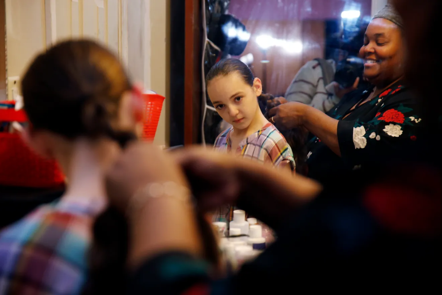 A woman is braiding a young girl's hair in front of a mirror, both smiling in a cozy room with art supplies nearby.
