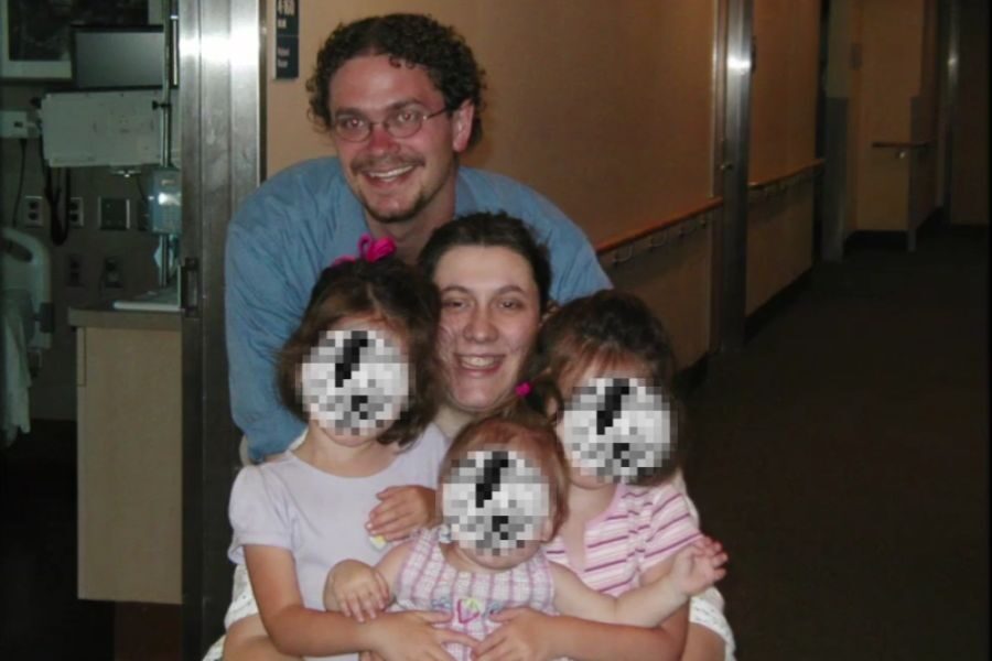A couple poses with three young girls whose faces are pixelated in what appears to be a hospital hallway. The man kneels behind the group smiling.